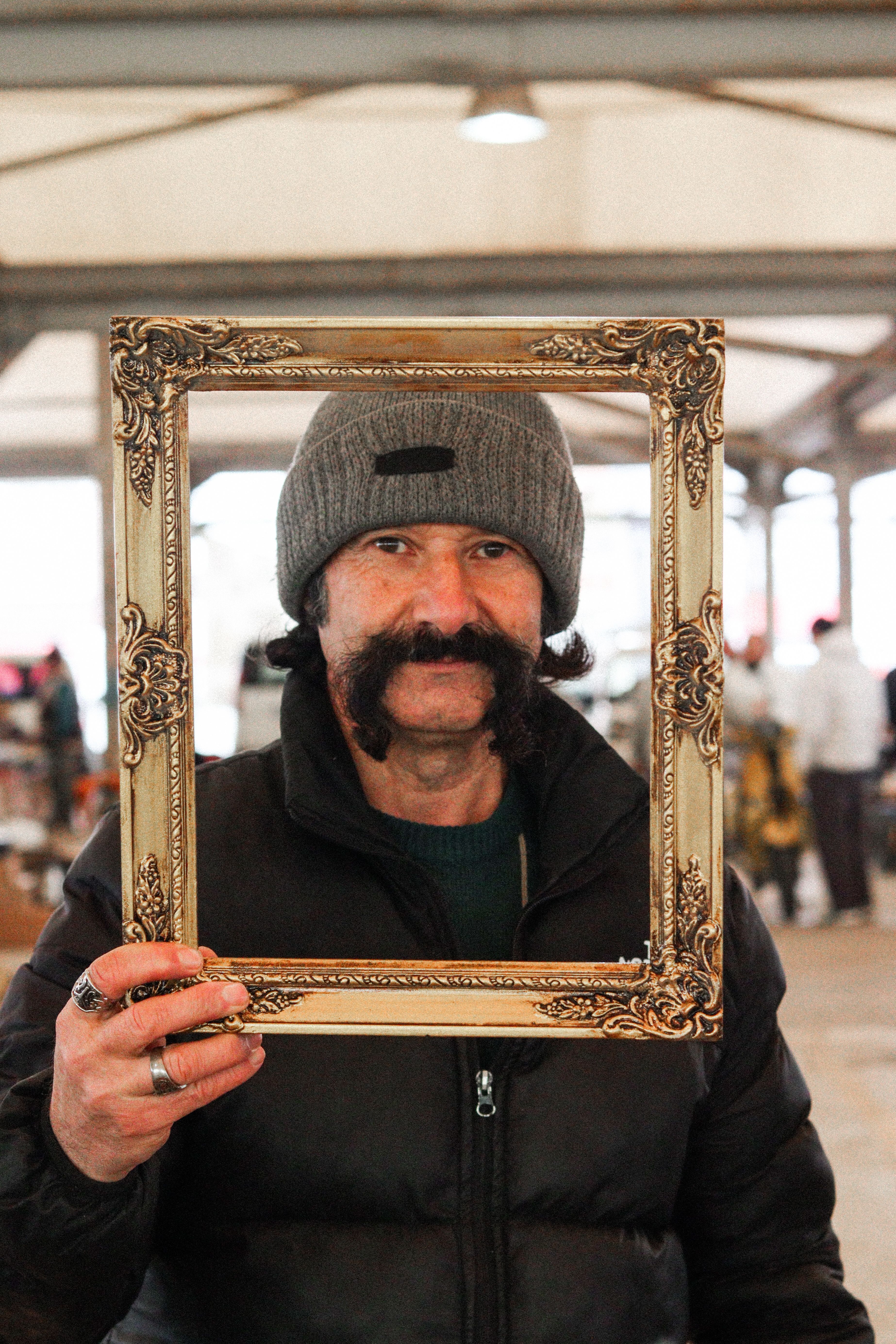 Man with a unique mustache holding a picture frame at an outdoor market.