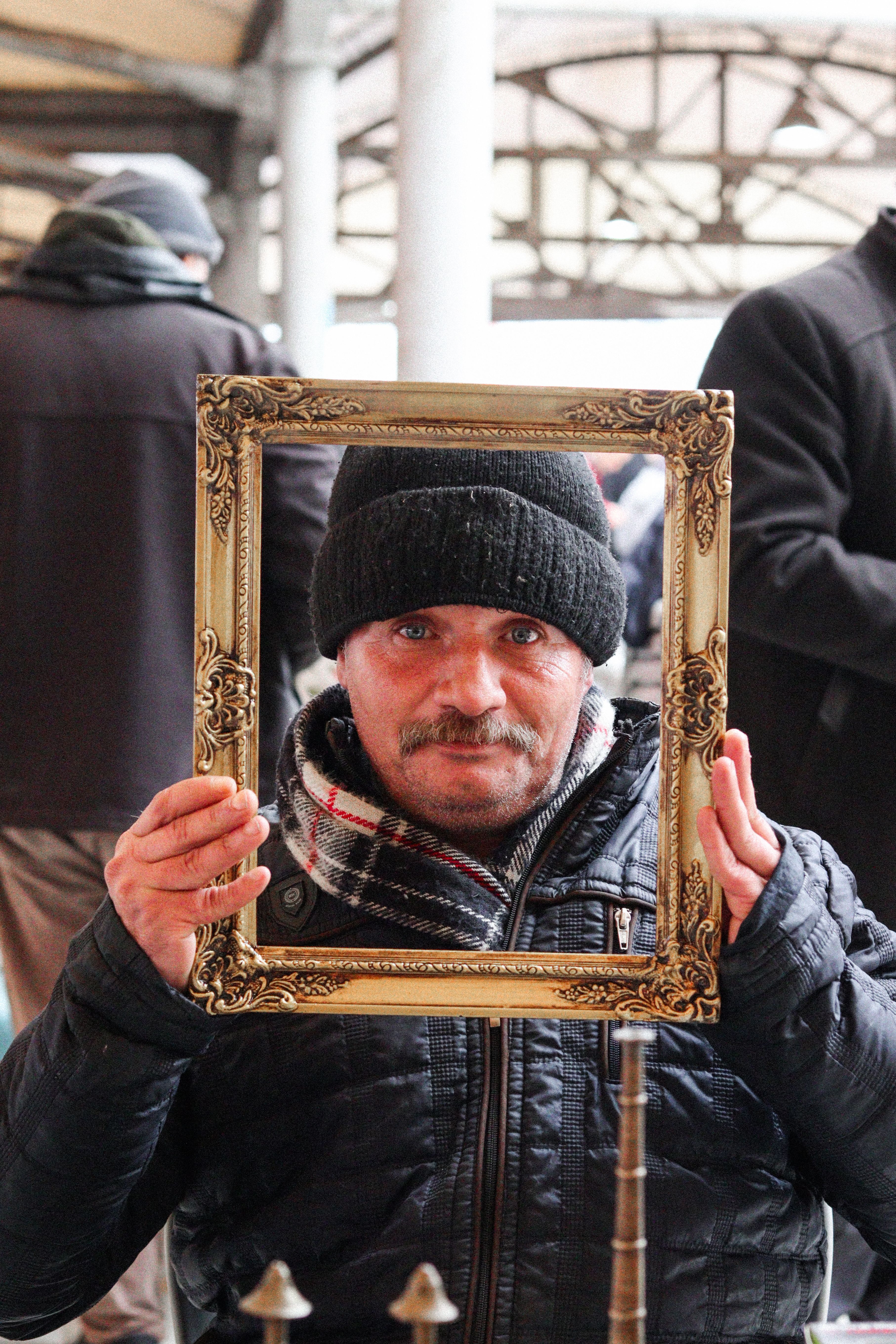 A man holding a decorative frame creates a unique portrait at a bustling market, wearing winter attire.