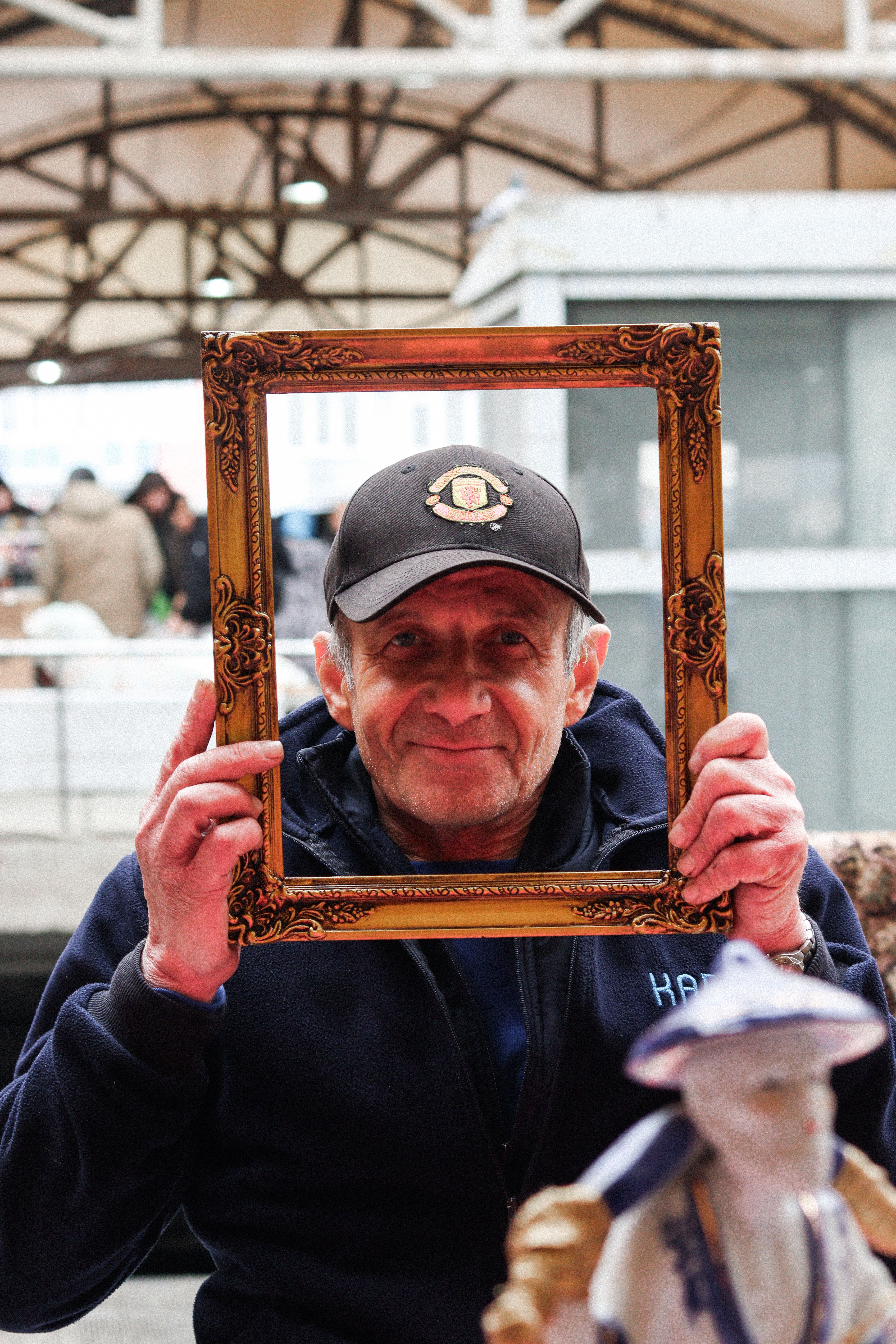 Elderly man with cap holding ornate frame at an outdoor market setting.