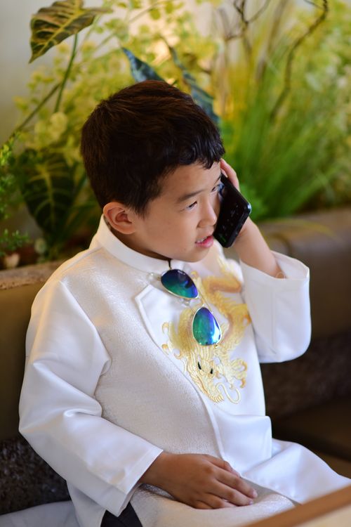 Free Child in a traditional white outfit talks on phone while seated, wearing sunglasses. Stock Photo