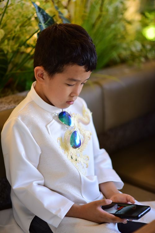 Free A young boy in white traditional attire sits indoors using a smartphone. Stock Photo