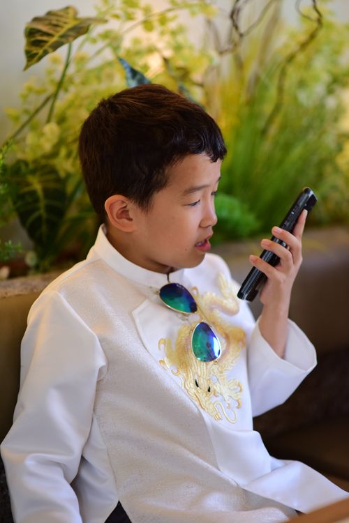 Free A young boy in a white traditional outfit sits indoors, engaging with a smartphone. Stock Photo