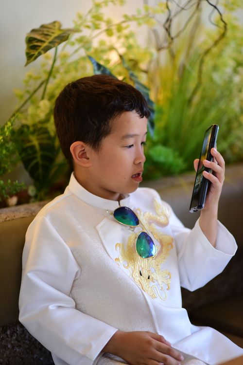Free A young boy in a white traditional outfit looks intently at a smartphone indoors. Stock Photo