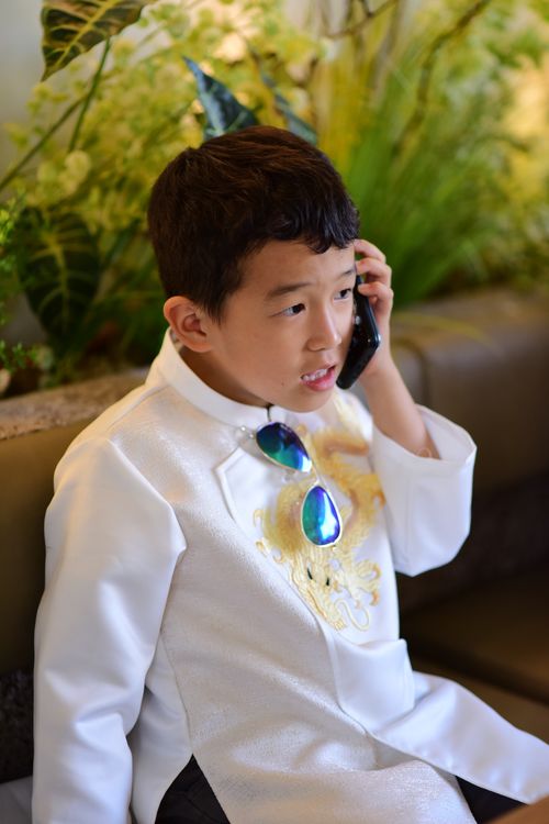 Free A young boy in traditional white clothing holds a phone, sitting indoors with plants in the background. Stock Photo