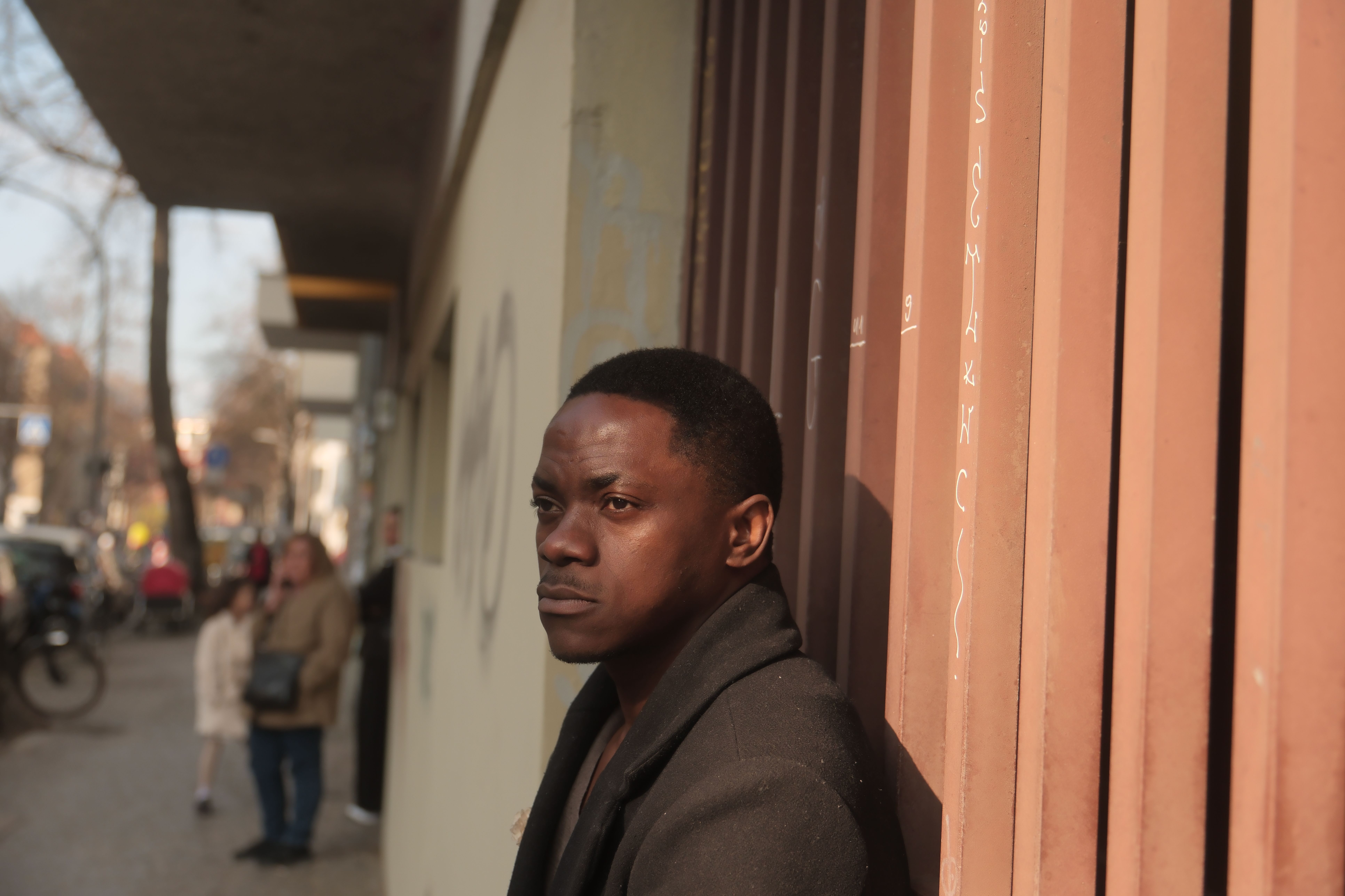 A man leans against a wall in a thoughtful pose on a sunny Berlin street.