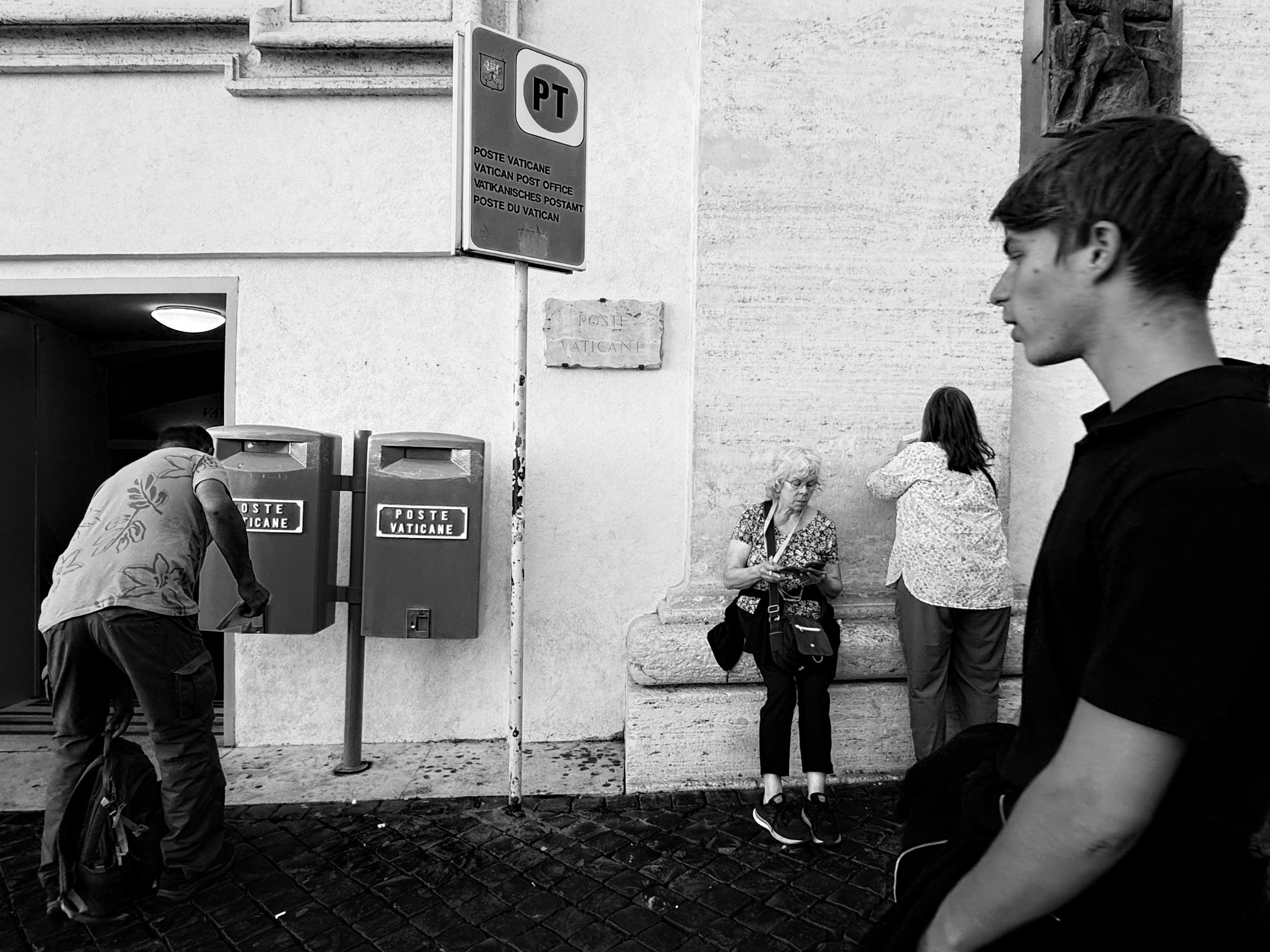 Black and white candid street scene at Vatican City post office with people capturing travel moments.