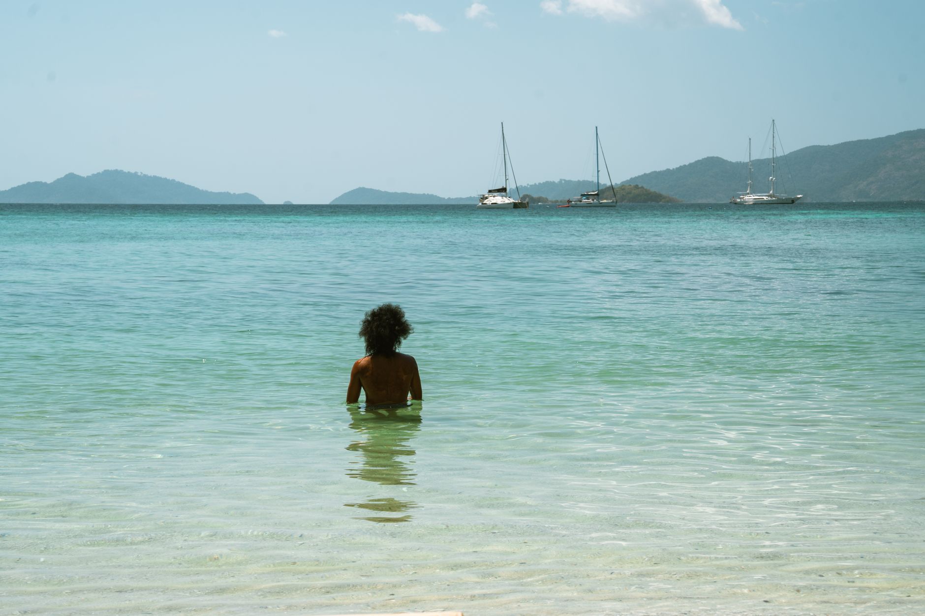 A lone individual relaxes in the clear ocean waters, facing distant anchored sailboats.