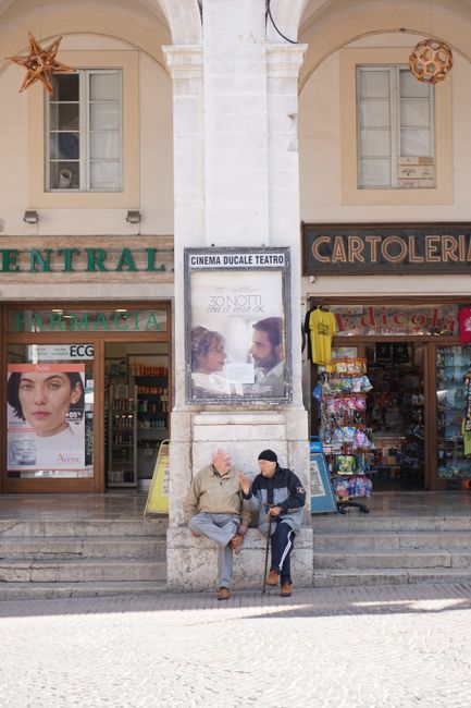 Two elderly men converse in a picturesque Italian arcade in Urbino's town center.