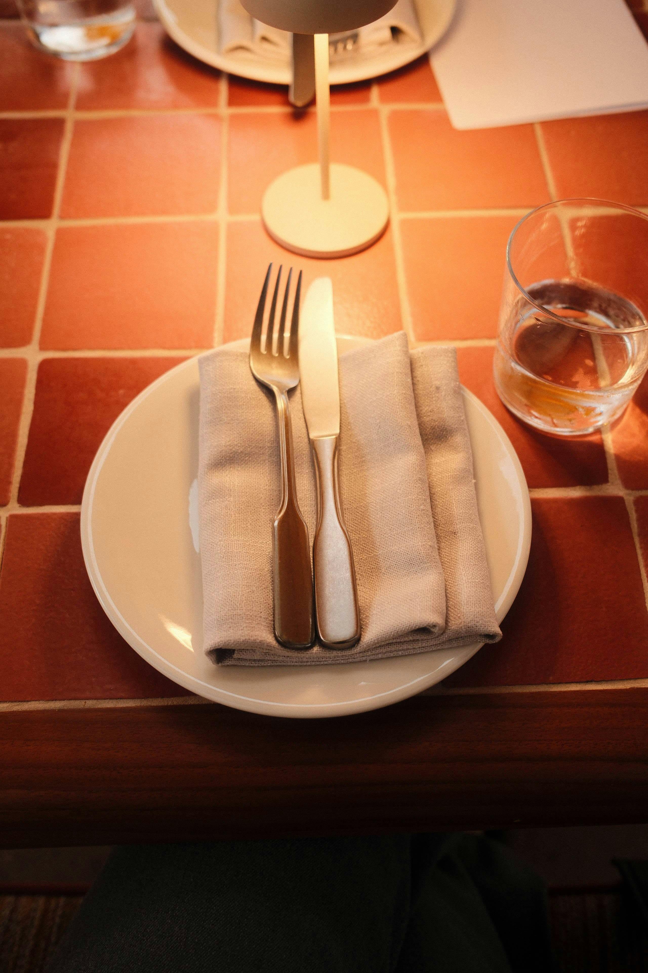 Fork and knife on napkin over plate on tiled table with glass of water.