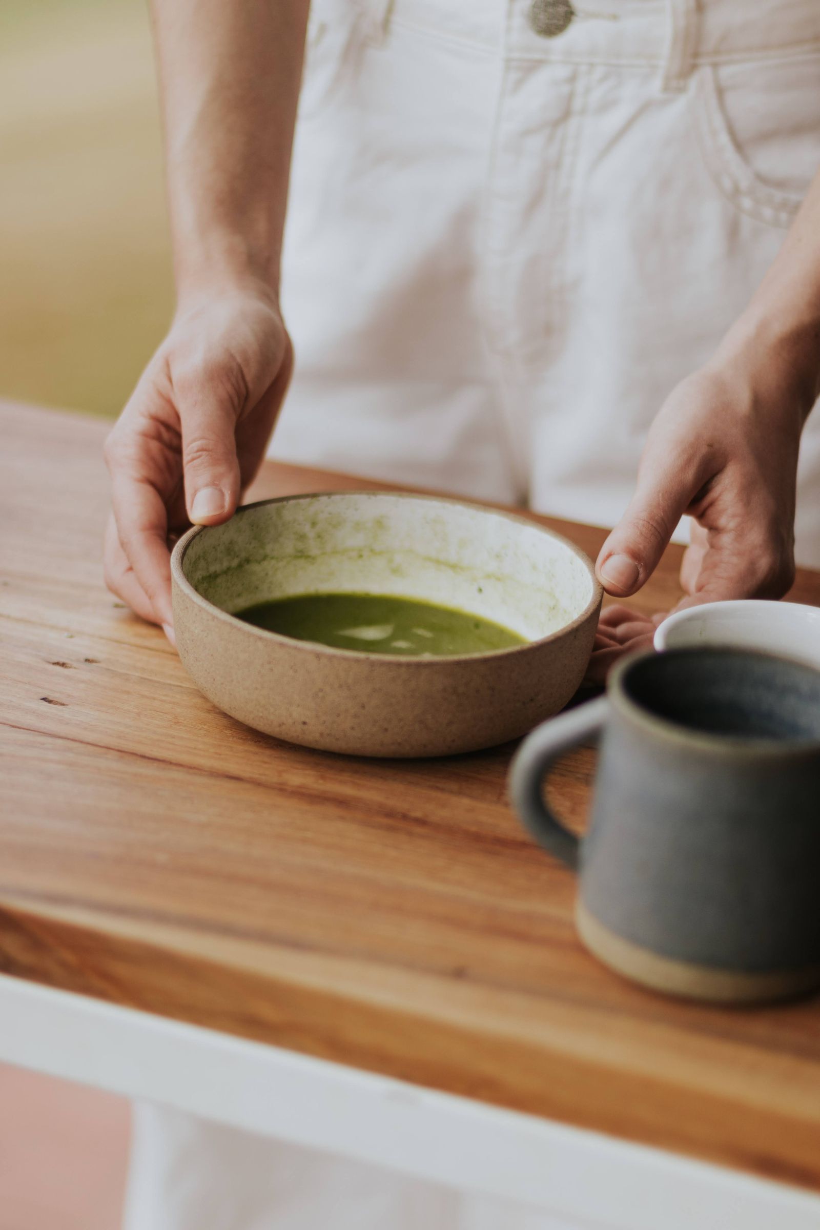 Person holding a ceramic bowl of matcha on a sunlit wooden table