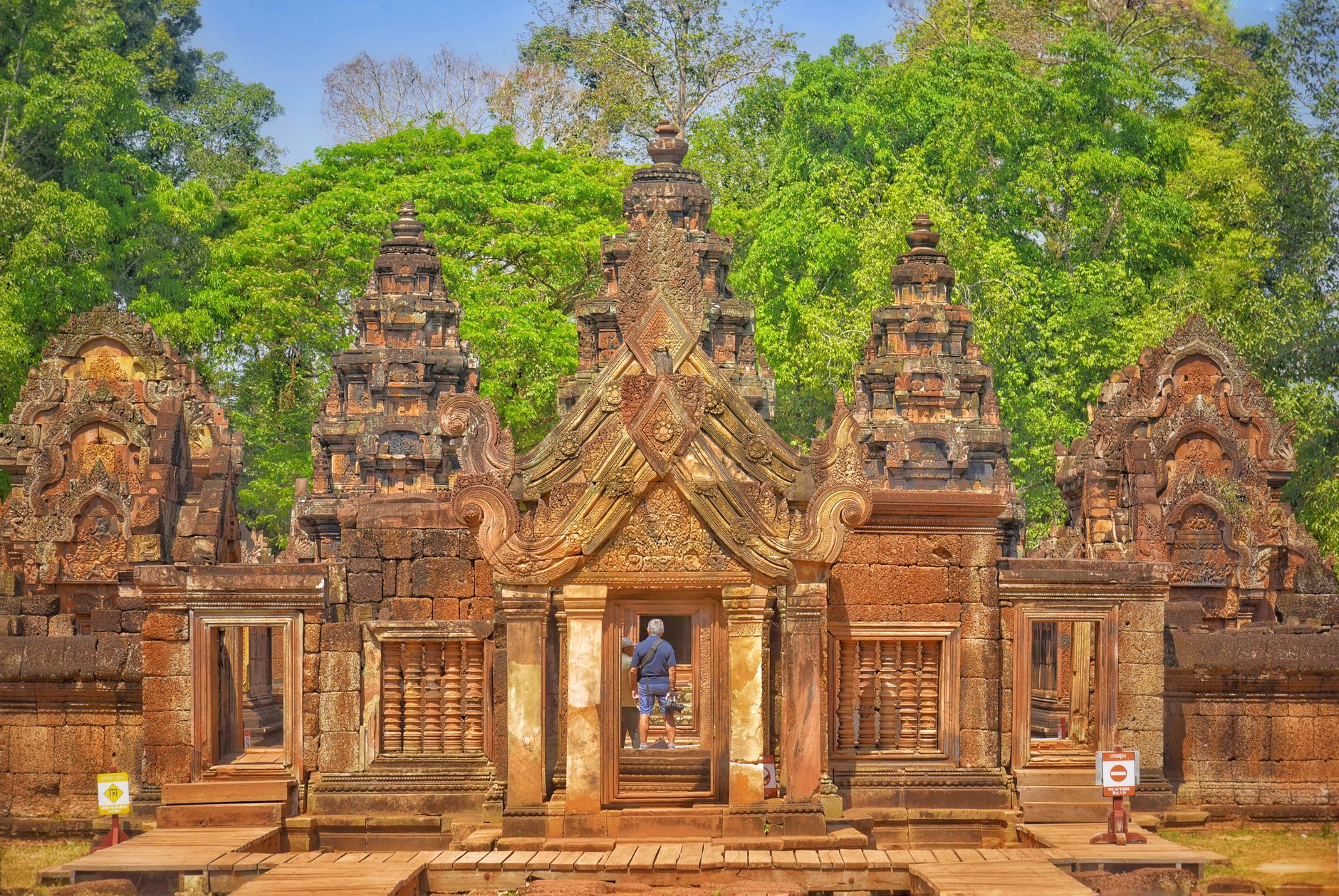 The pink-and-red sandstone shrines of Banteay Srei with their ornate carved pediments, a visitor standing in the central doorway