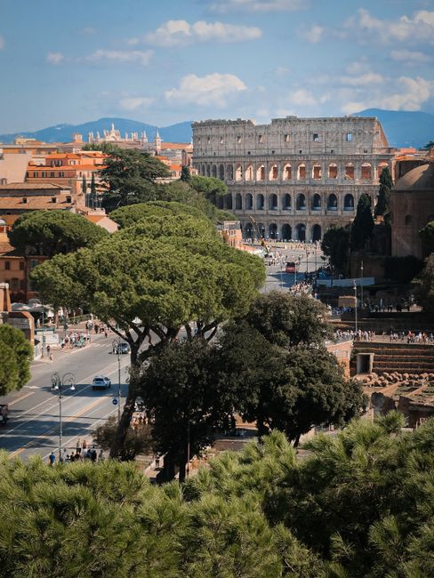 Aerial shot of the iconic Colosseum in Rome, Italy, surrounded by lush greenery under a bright sky.