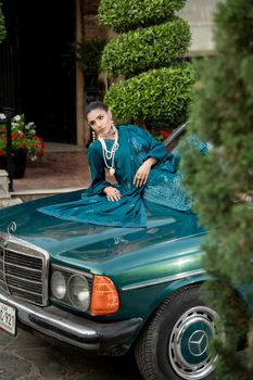 A fashion model wearing a blue outfit poses elegantly on a classic car.