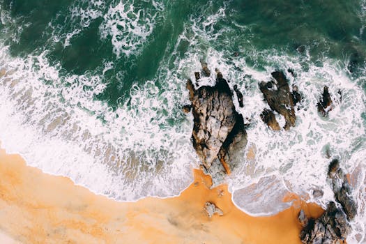 Aerial shot captures waves crashing on rocky shore with sandy beach.