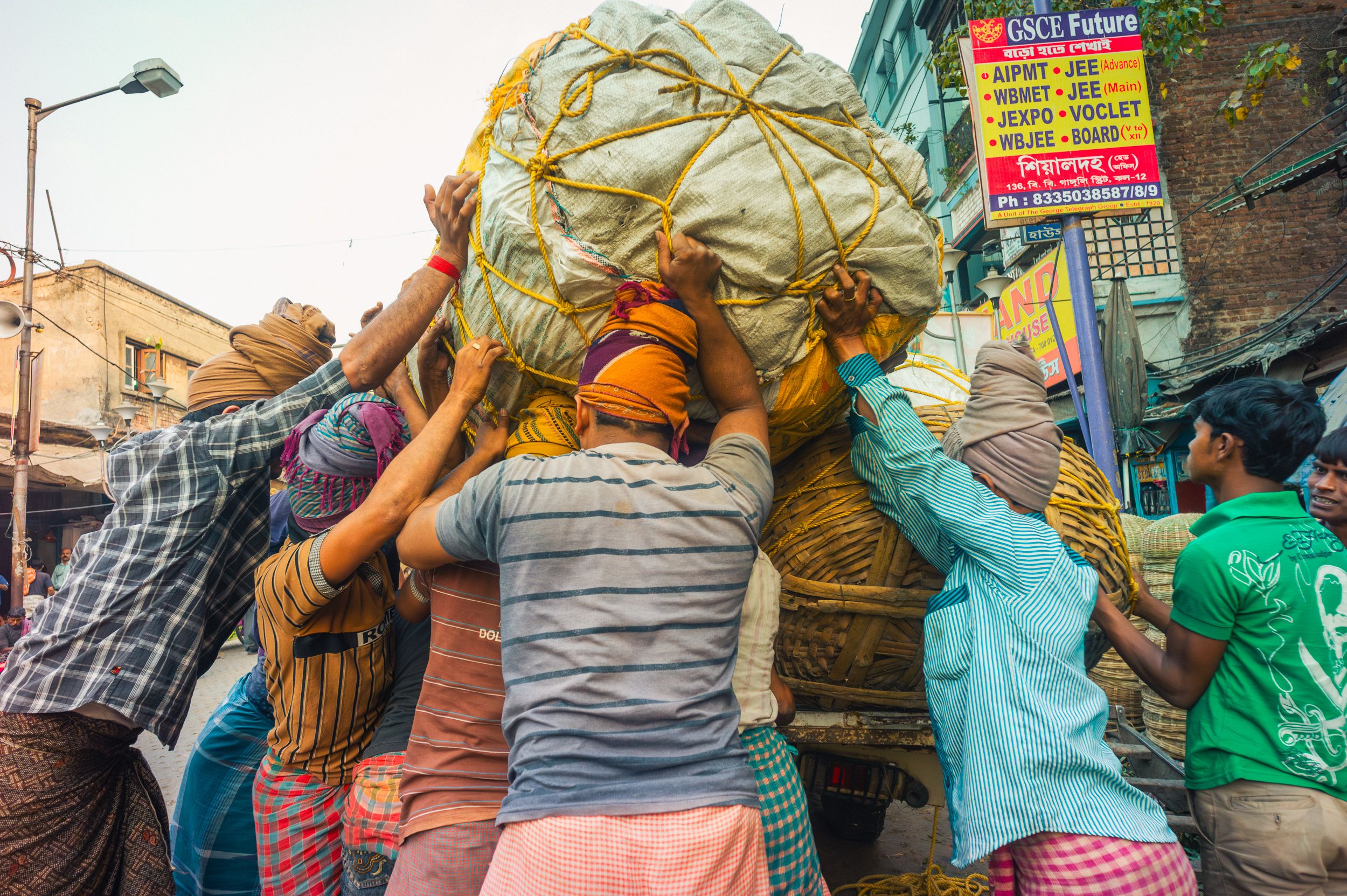 Group of laborers lifting heavy cargo in Kolkata market street, showcasing teamwork and local urban life.