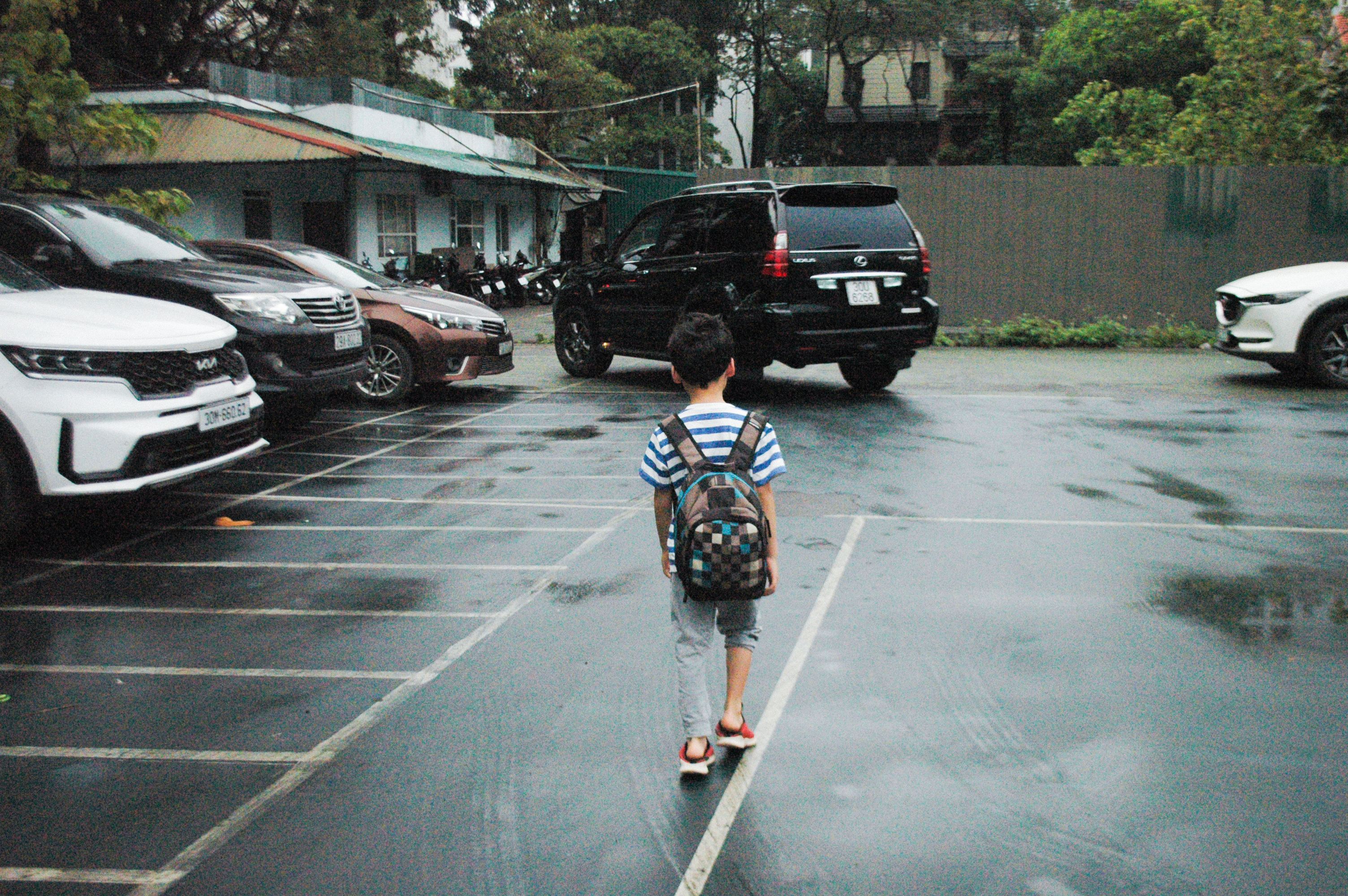 A young boy with a backpack walks through a parking lot, surrounded by cars on a rainy day.