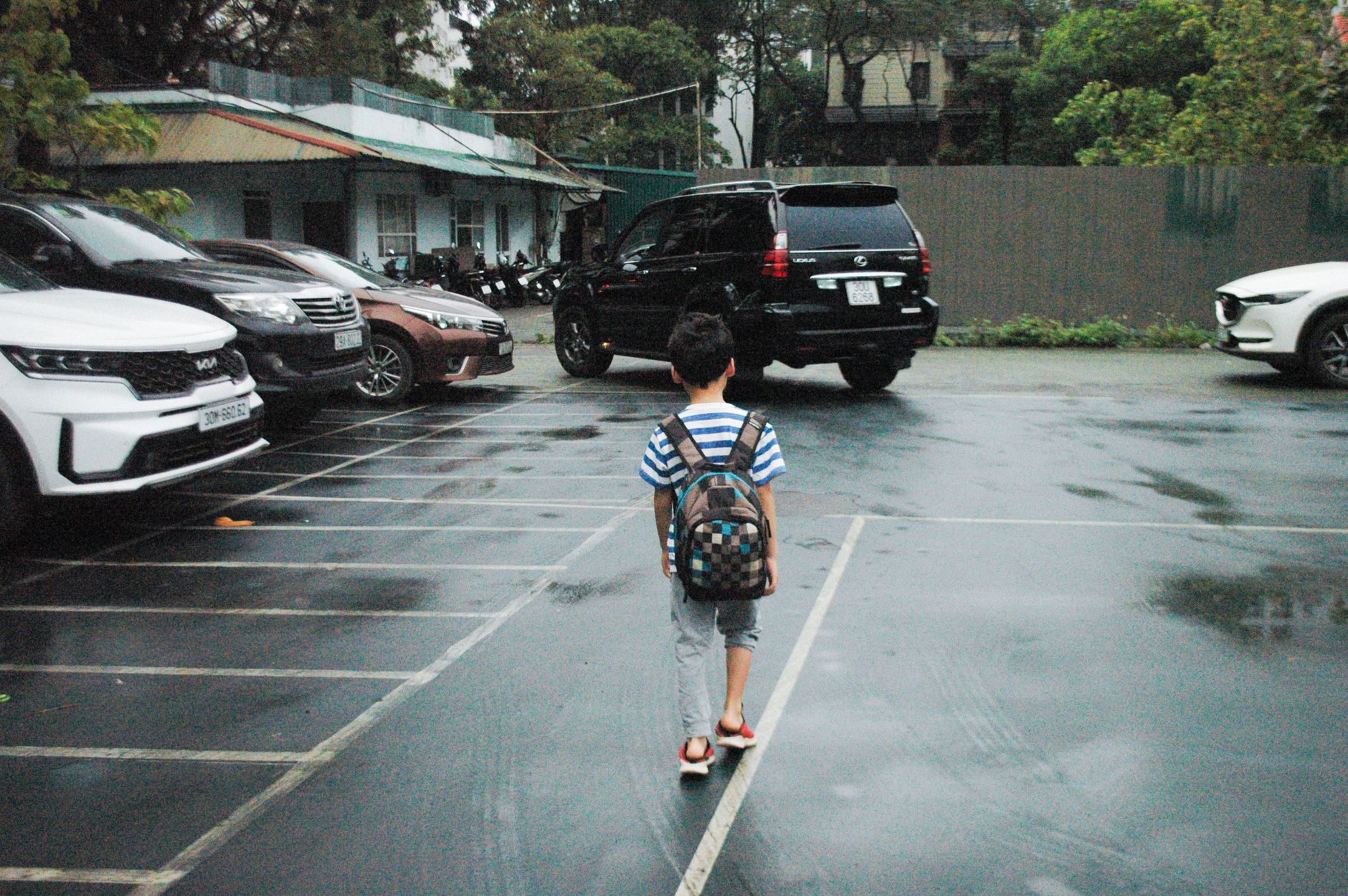 A young boy with a backpack walks through a parking lot, surrounded by cars on a rainy day.