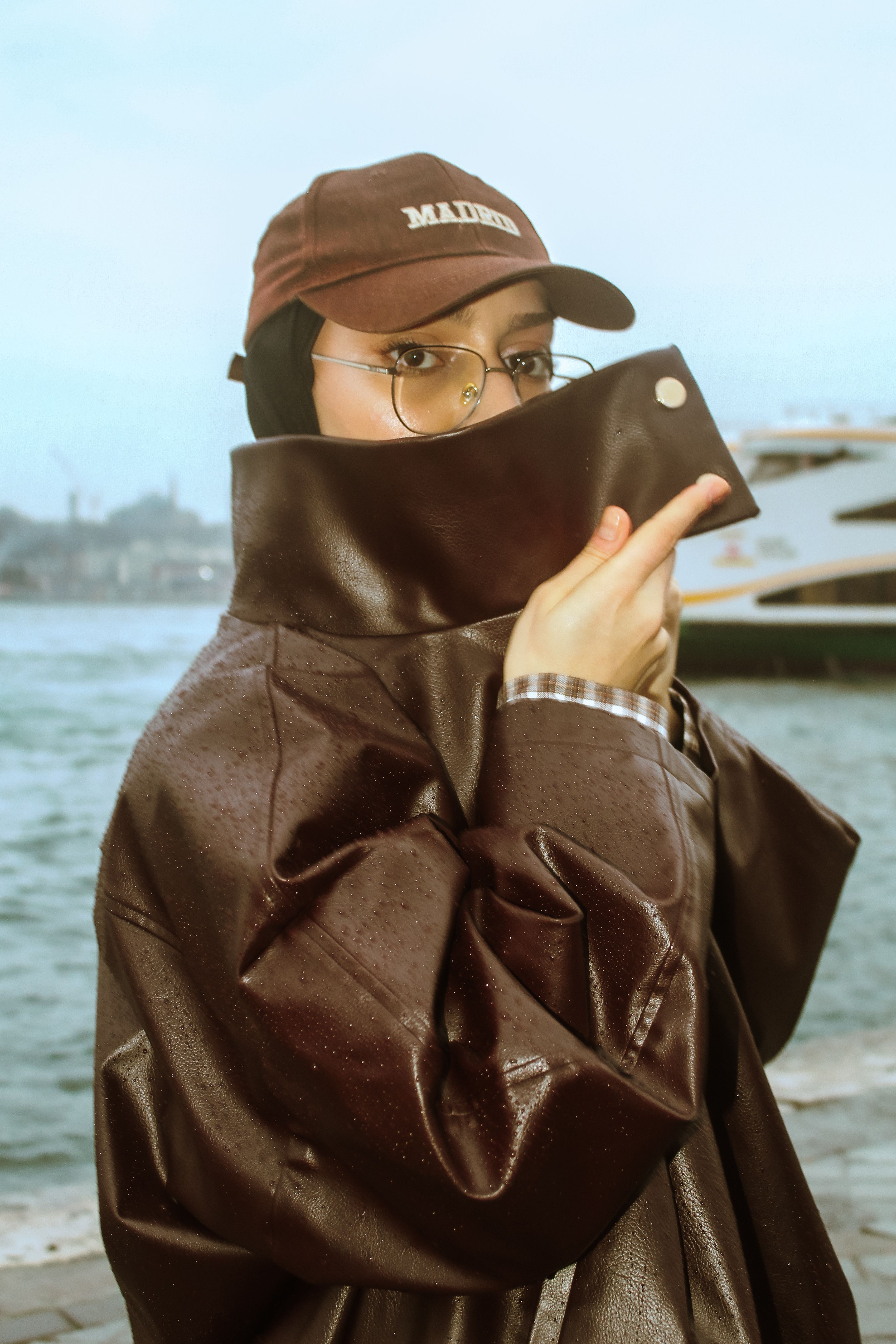 A stylish woman in a brown jacket and cap poses by the waterfront, exuding urban fashion vibes.