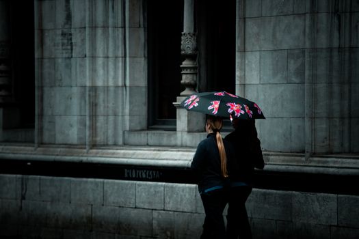 Two adults with a floral umbrella walk by a historic stone facade in rainy Ireland.