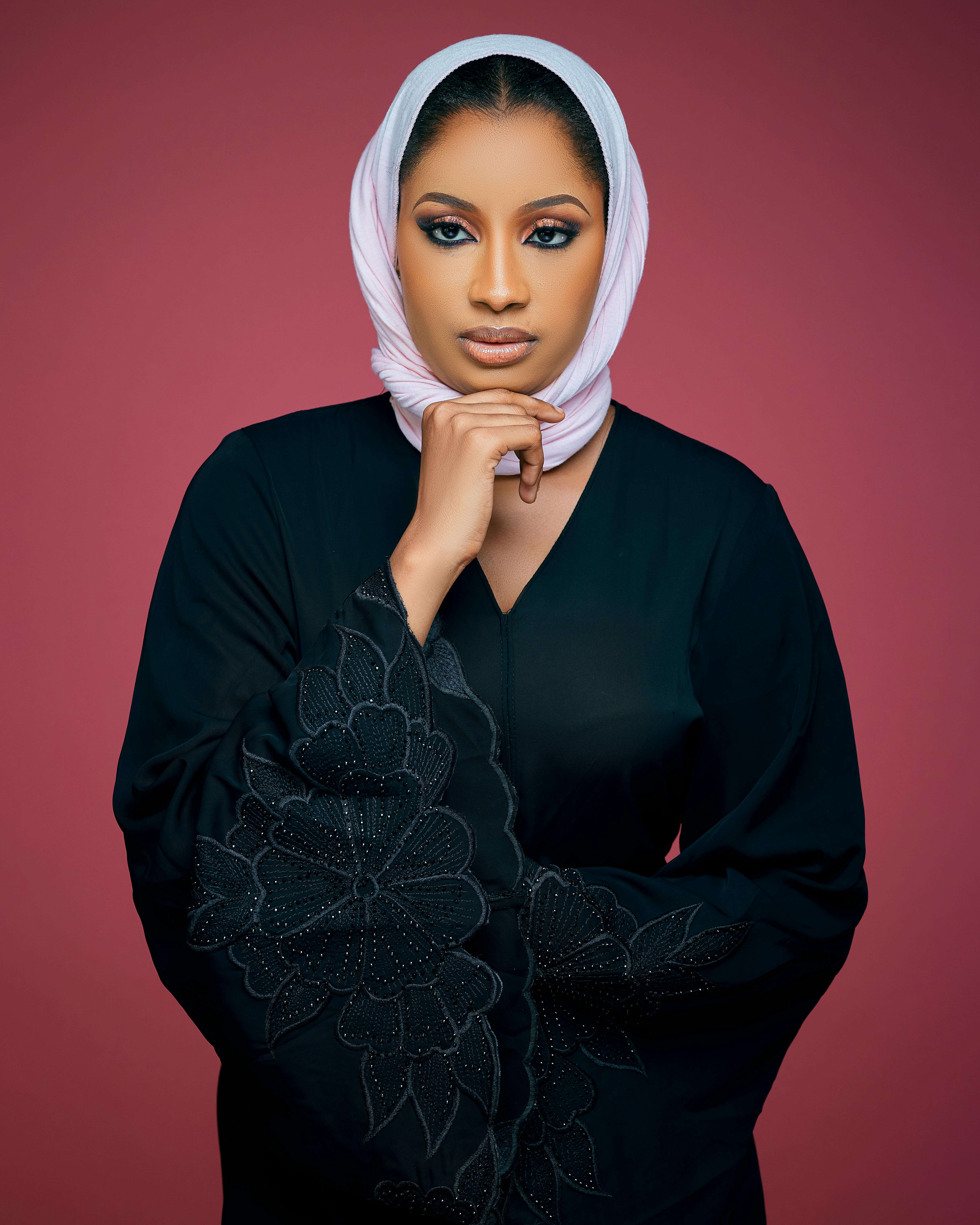 Portrait of a woman wearing a hijab with intricate embroidery, set against a red backdrop.