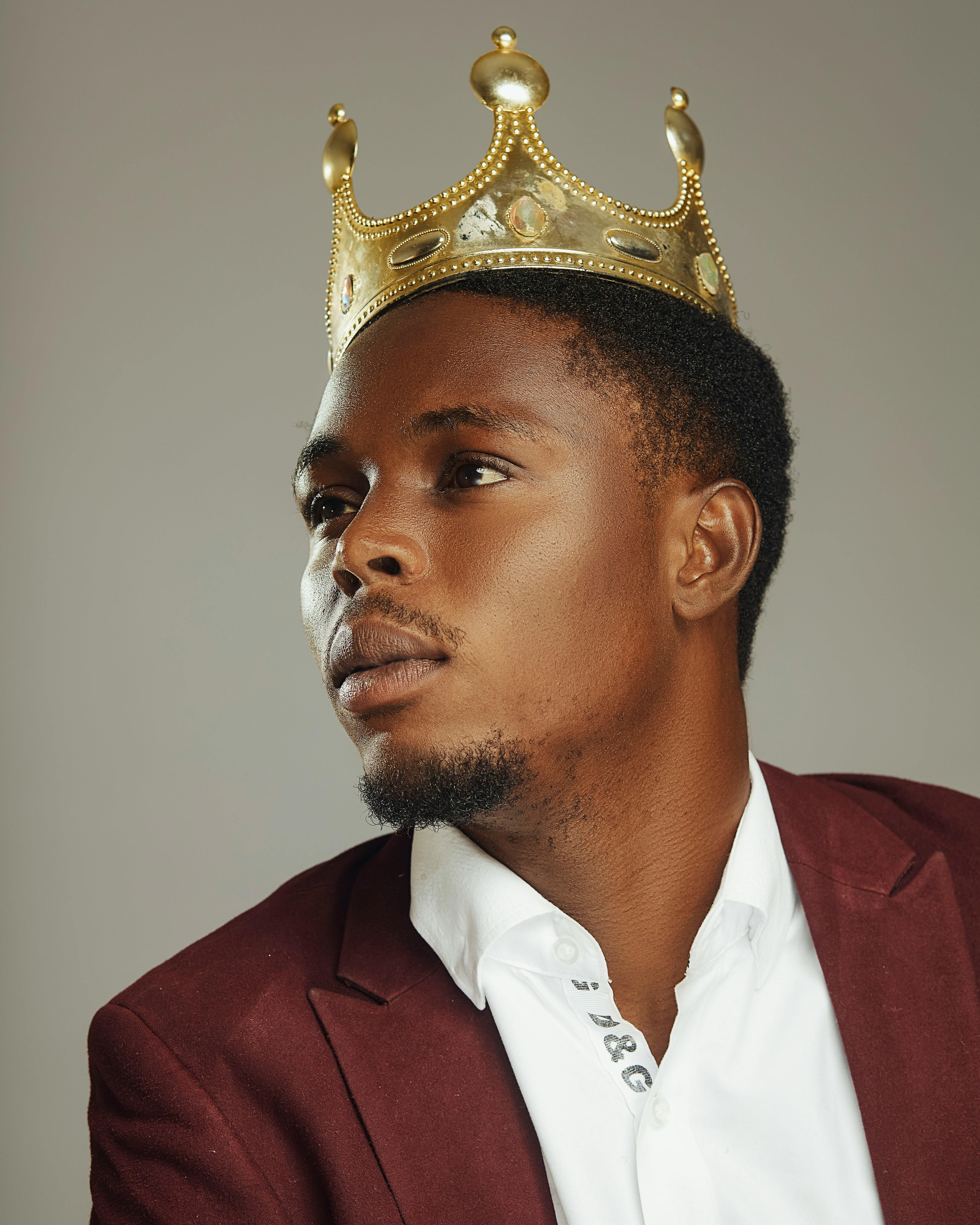 Regal portrait of a young man in a suit with a gold crown against a neutral background.