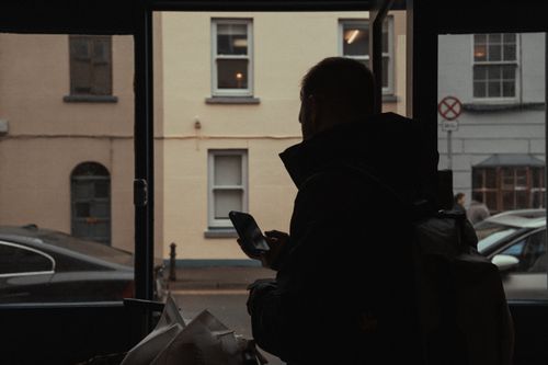 Free Silhouette of a person using a smartphone near a window, overlooking a street view. Stock Photo