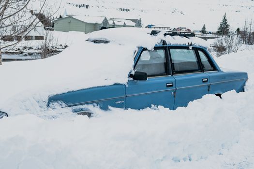 A blue vintage car sits buried in snow during a winter day outdoors.