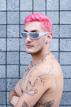 Stylish shirtless man with tattoos, pink hair, and sunglasses against a tiled wall.