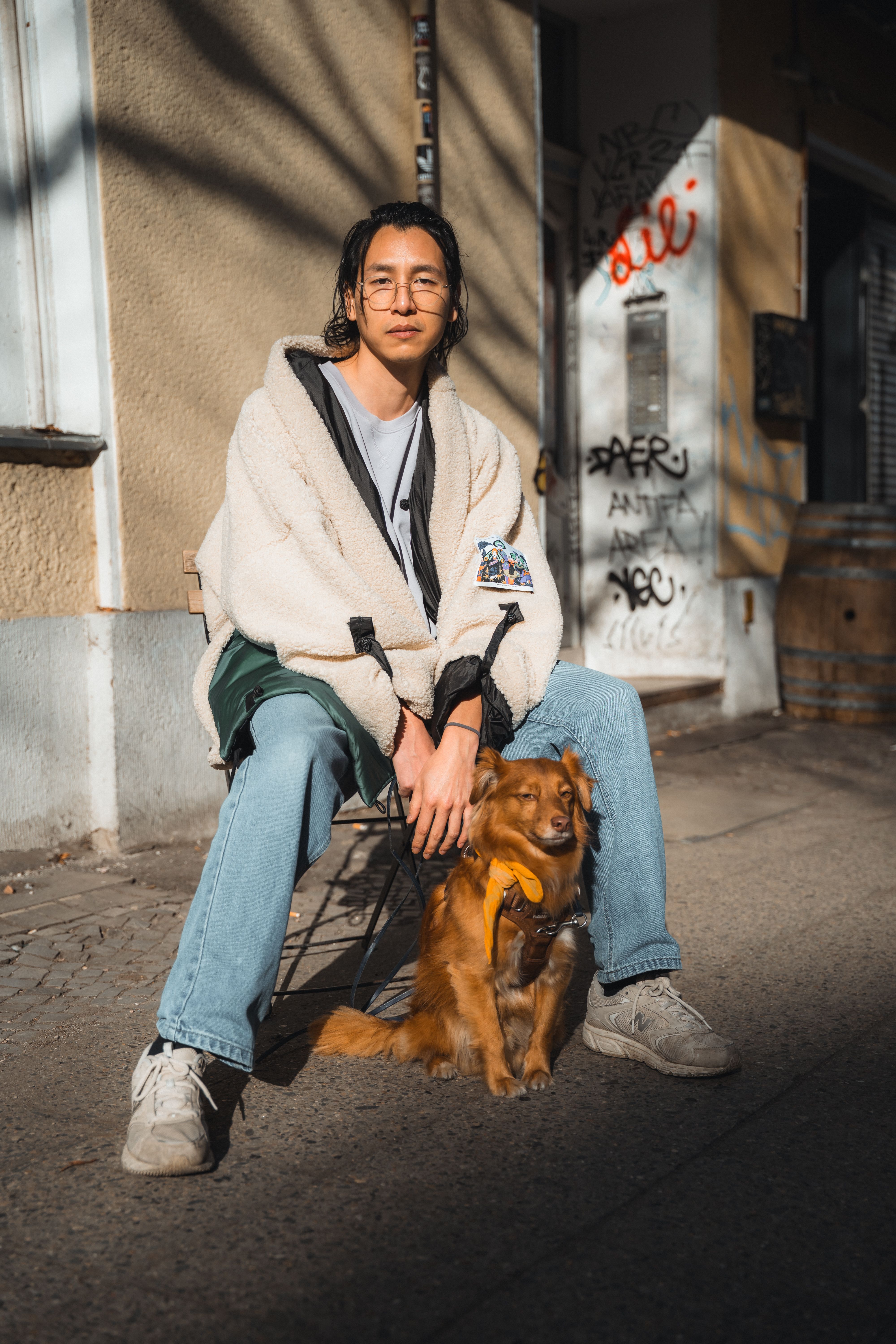 A young adult sits on a street in Berlin with a small dog. Urban setting with graffiti in the background.