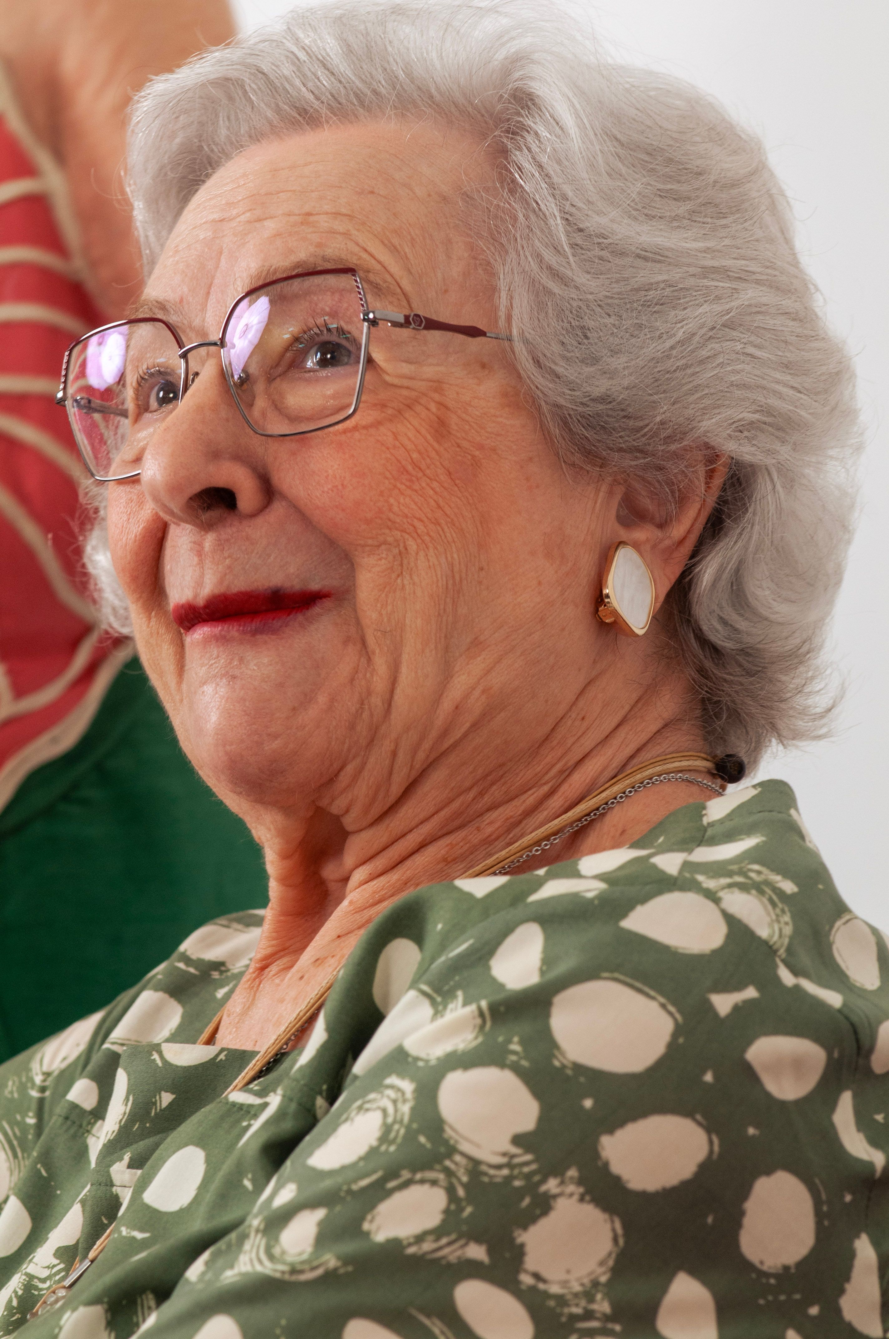Smiling senior woman wearing glasses and earrings in a green polka dot dress, exuding elegance.