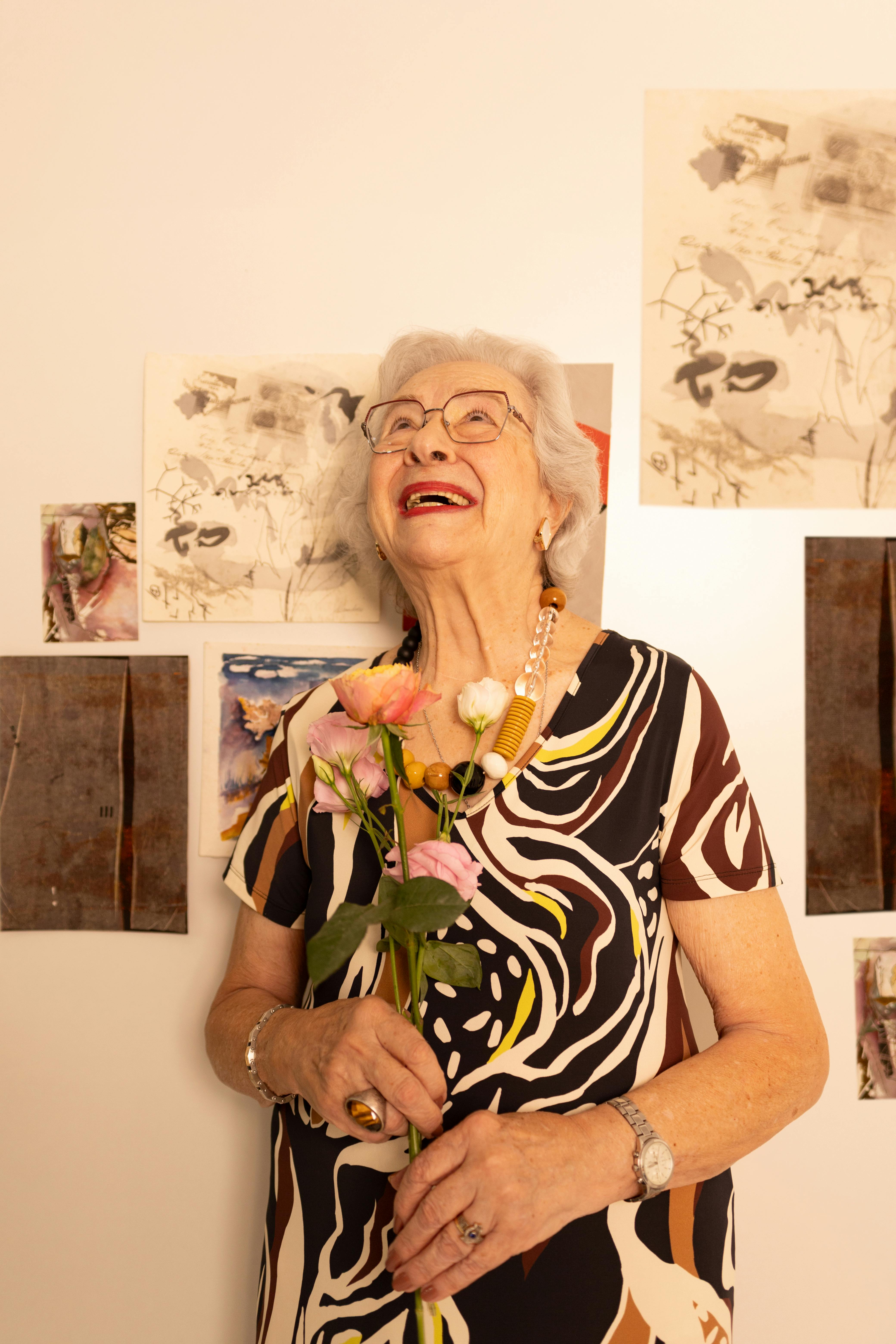 Elderly woman smiling warmly, holding flowers in front of abstract art indoors.