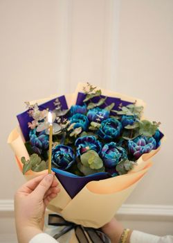 A vibrant bouquet of blue peonies with eucalyptus, held by a hand with a lit candle.