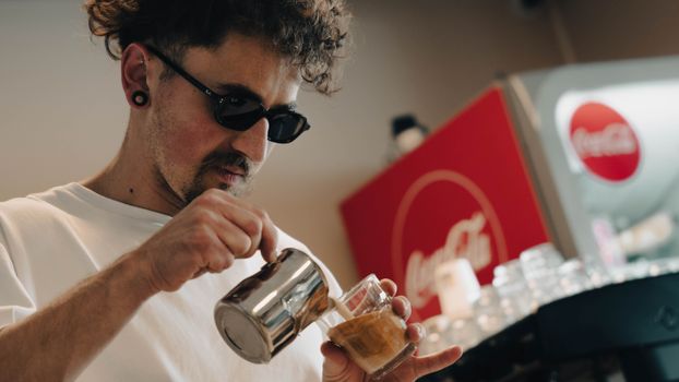 Barista with glasses skillfully pours milk into espresso, creating latte art in a cafe.