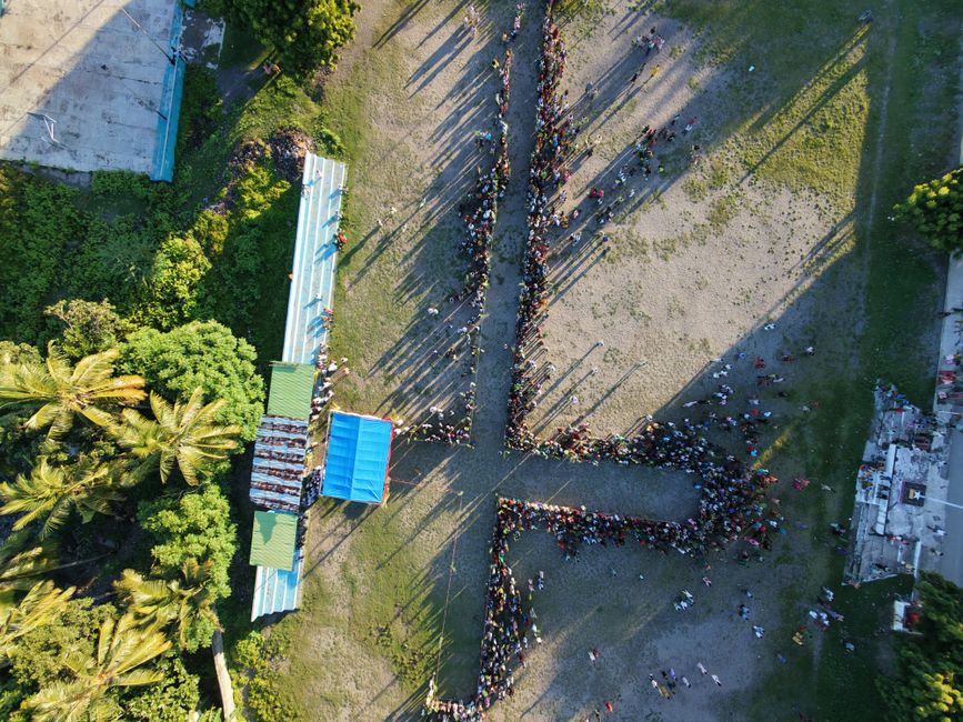 Aerial shot of a large outdoor gathering with people forming a pattern, surrounded by tropical greenery.
