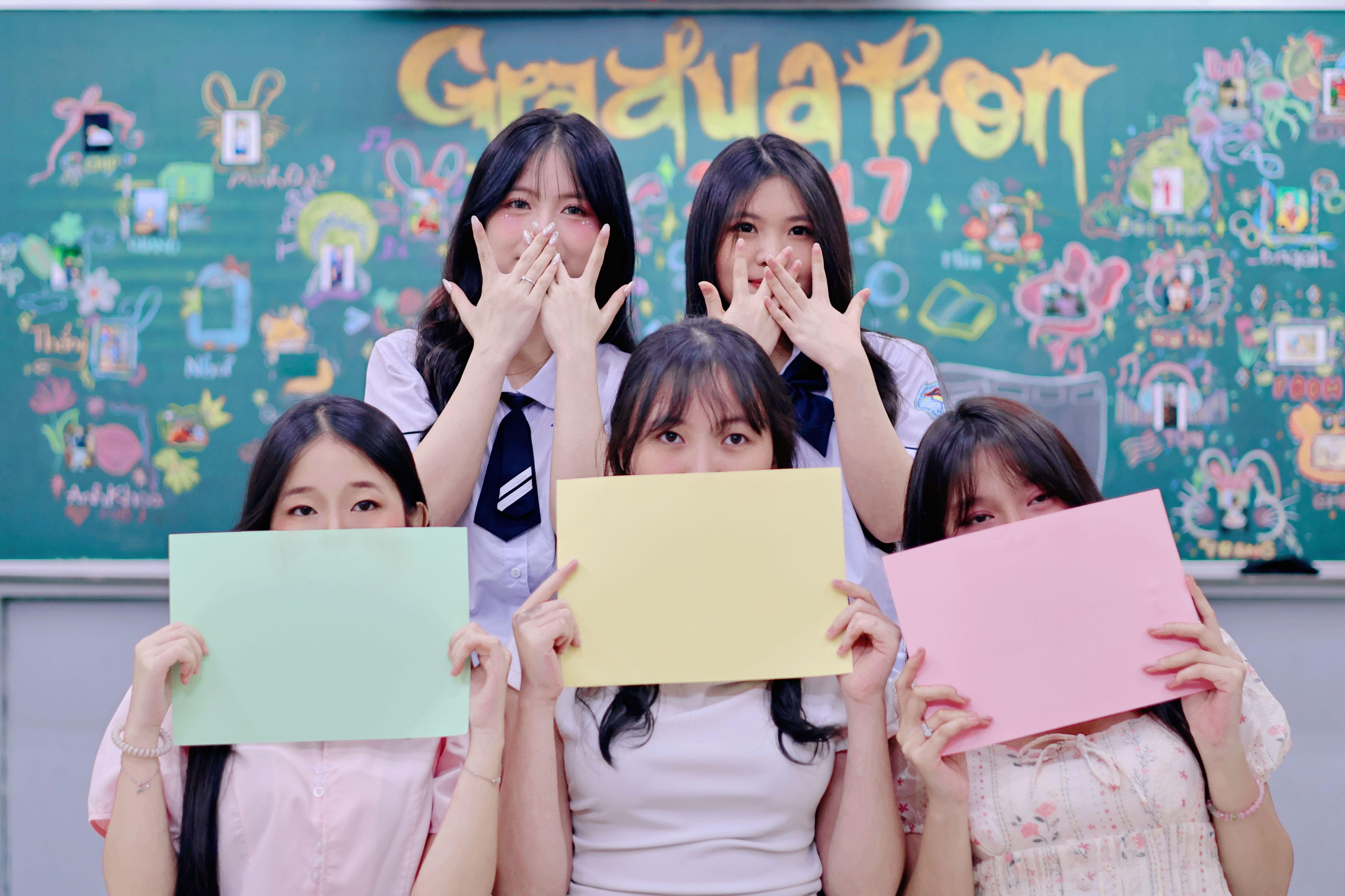 Lima siswa memegang kertas warna-warni di depan papan tulis berhias dalam perayaan kelulusan.