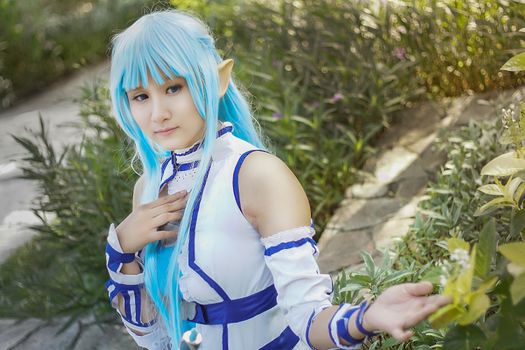 A whimsical cosplay character with blue hair poses in a lush Surabaya garden, capturing fantasy allure.