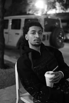 A young man with a coffee cup sits outdoors at night, with a parked SUV in the background.