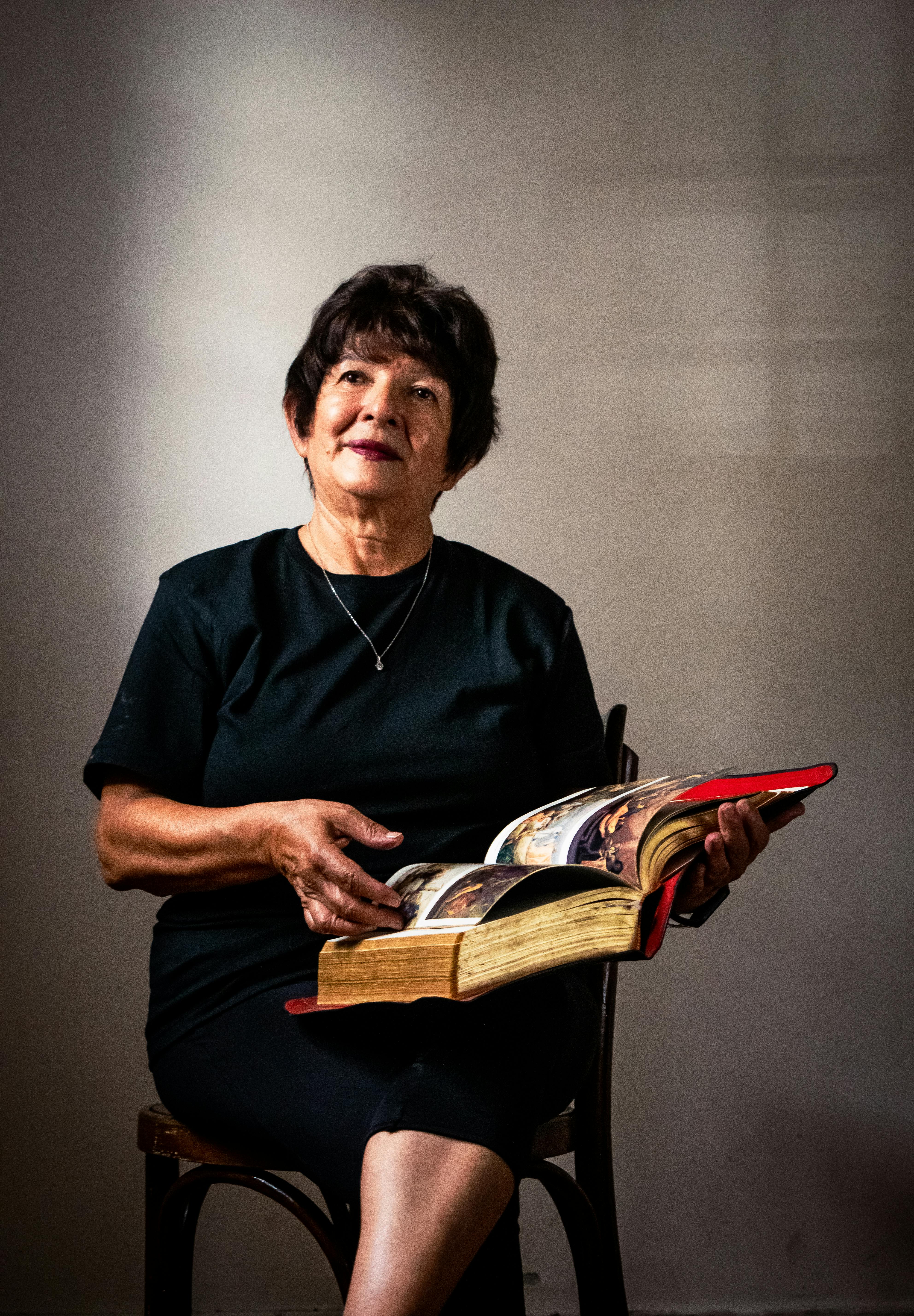 Elderly woman seated indoors, focused on reading a large book with thoughtful expression.