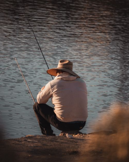 Free A serene scene of a man fishing by a tranquil lake during a warm sunset. Stock Photo