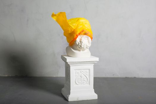 A classical marble bust adorned with a vibrant yellow plastic bag, creating a striking visual statement.
