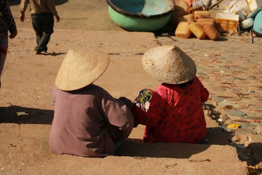 Two Vietnamese locals in conical hats sitting on a cobbled path, with colorful containers nearby.