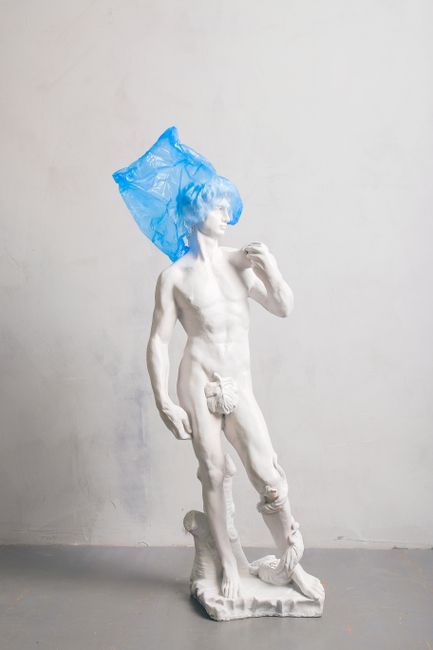 A classical nude sculpture adorned with a blue plastic bag, blending art with environmental themes.