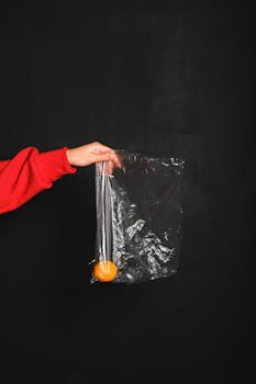 一只戴着红袖子的手，拿着一个装着橙子的透明塑料袋，背景是深色的