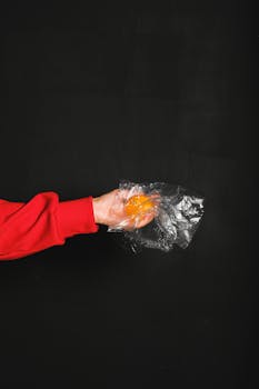 一只戴着红袖套的手，在深色背景中握着一个塑料包裹的橙子，象征着对环境的担忧