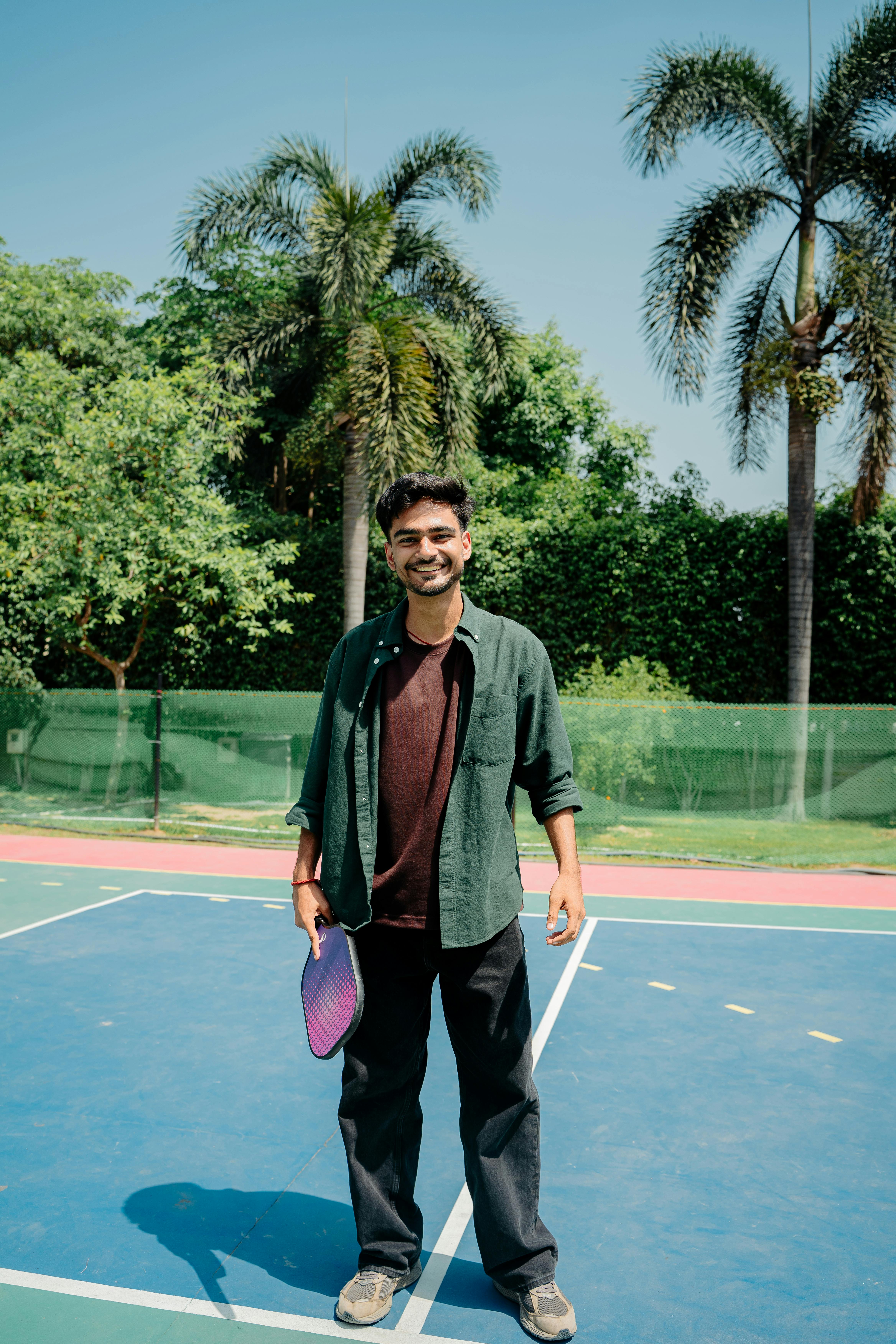 A cheerful young man standing on a pickleball court with a paddle, surrounded by lush greenery.