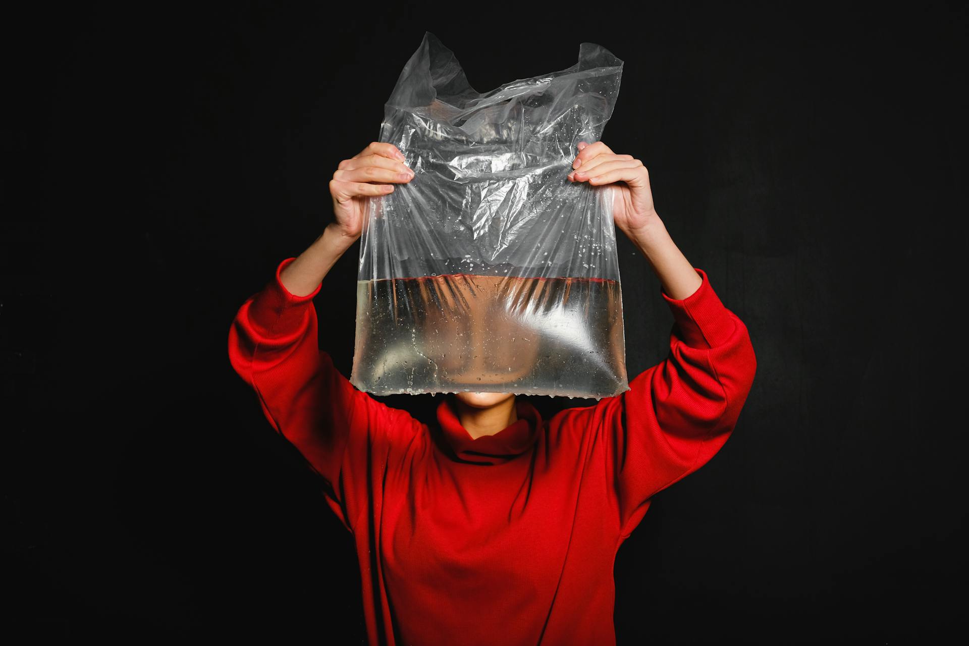 A person in a red sweater holds a water-filled plastic bag in front of their face against a dark studio background.