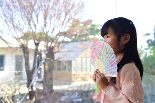 Free A young girl in a traditional outfit holding a decorative fan and looking out a window. Stock Photo
