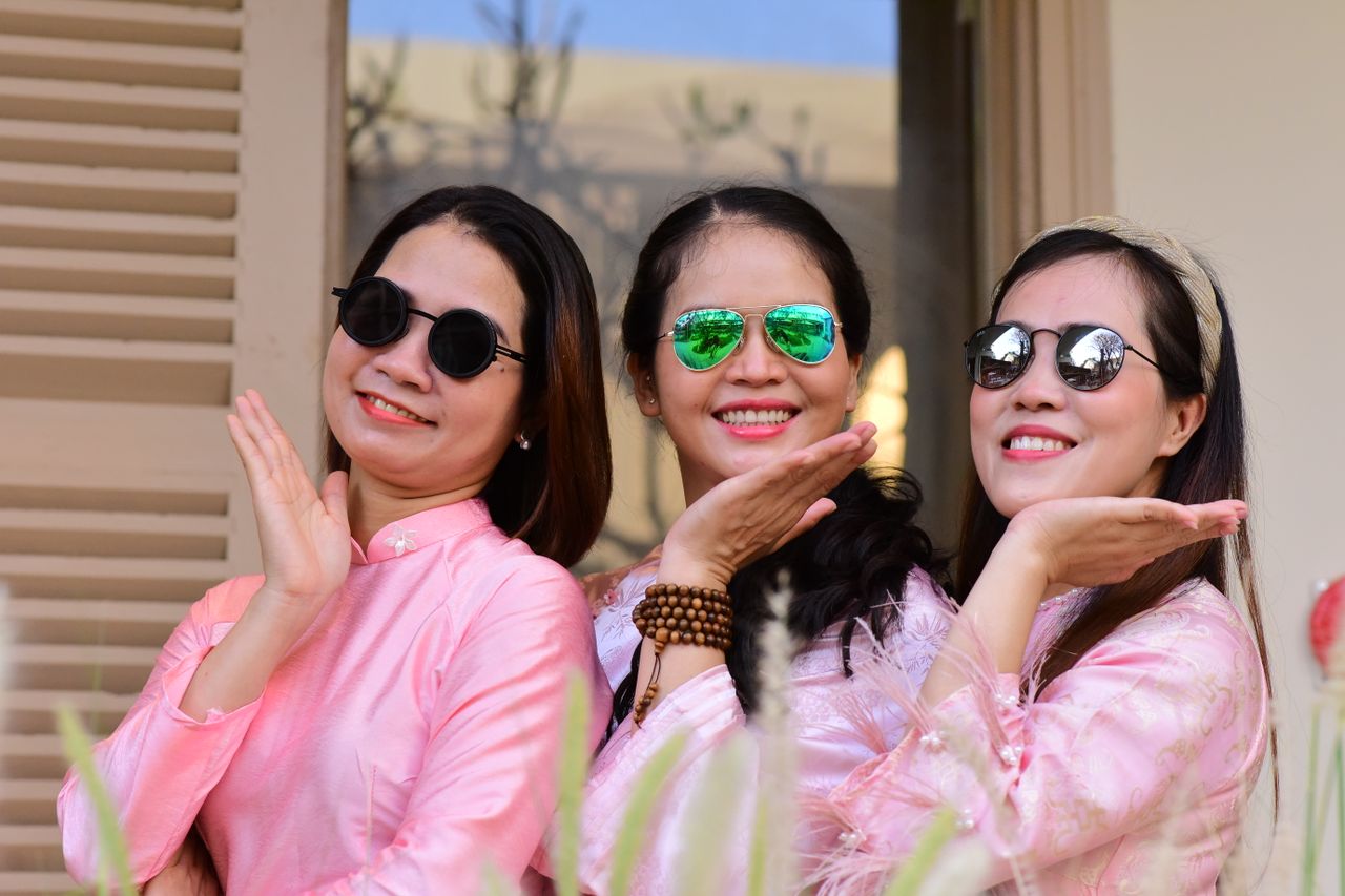 three women wearing sunglasses and pink ao dai posing happily outdoors