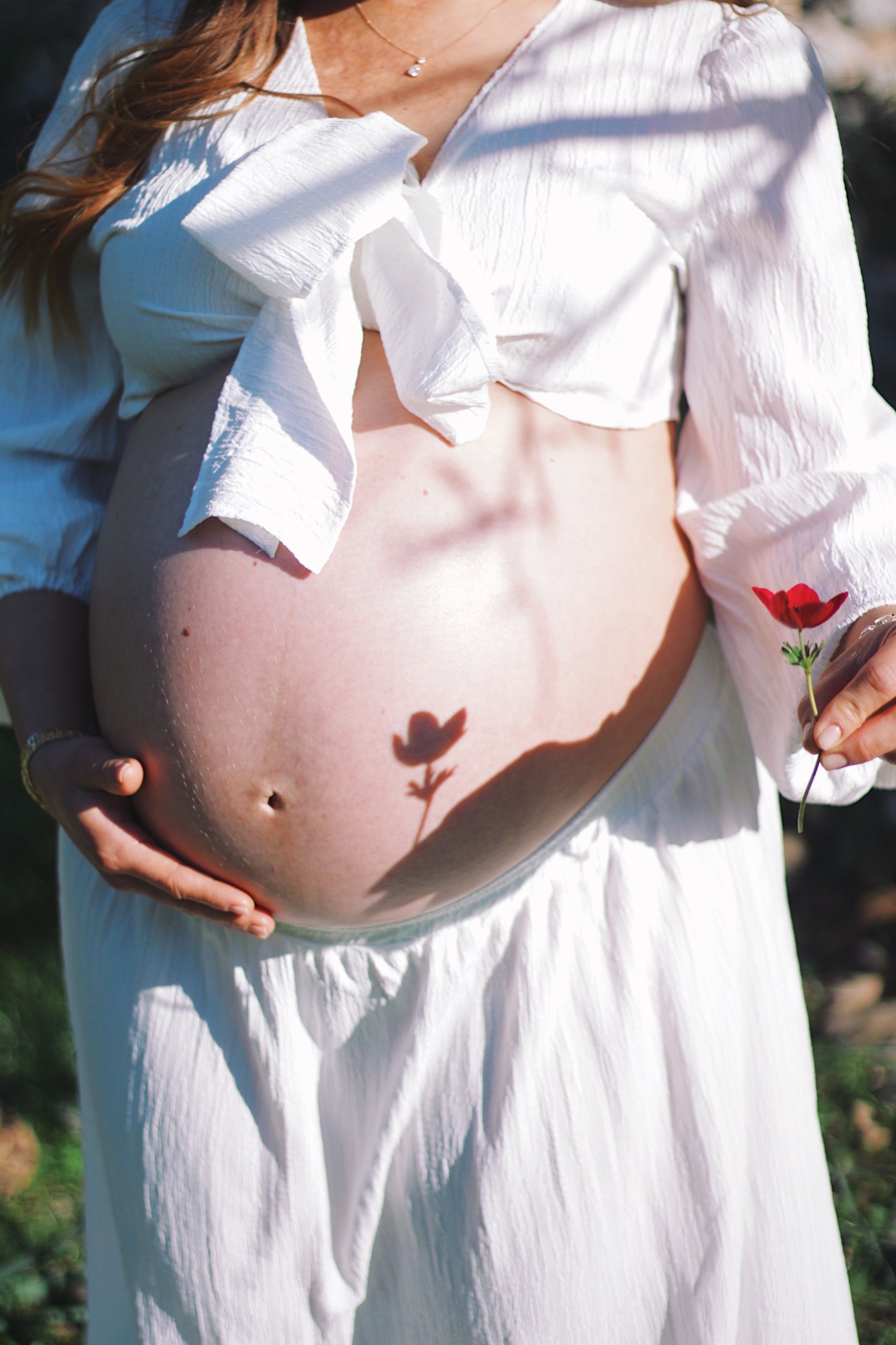 Pregnant woman in white dress holding a red flower with its shadow on her belly, outdoors in Bodrum.