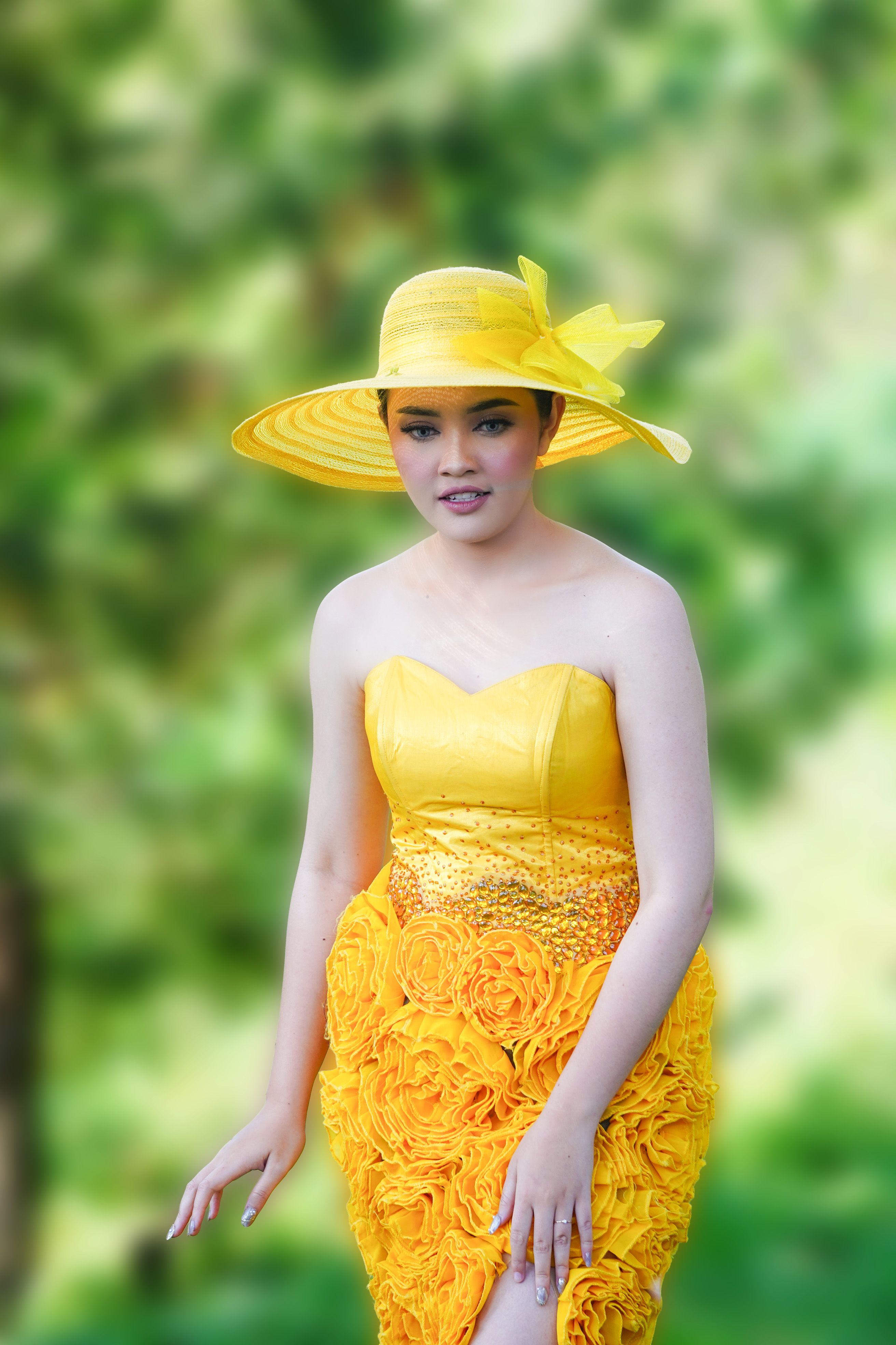 A woman in a bright yellow dress and matching hat poses outdoors amidst lush greenery, exuding elegance and style.